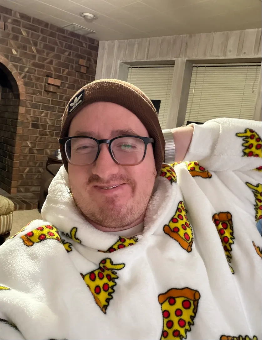 Cam, wearing glasses and a beanie, wrapped in a pizza-print blanket at home.